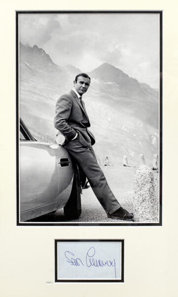 A photograph of Sean Connery as 007 with the Goldfinger Aston Martin DB5 in the Furka Pass, mounted with an autograph